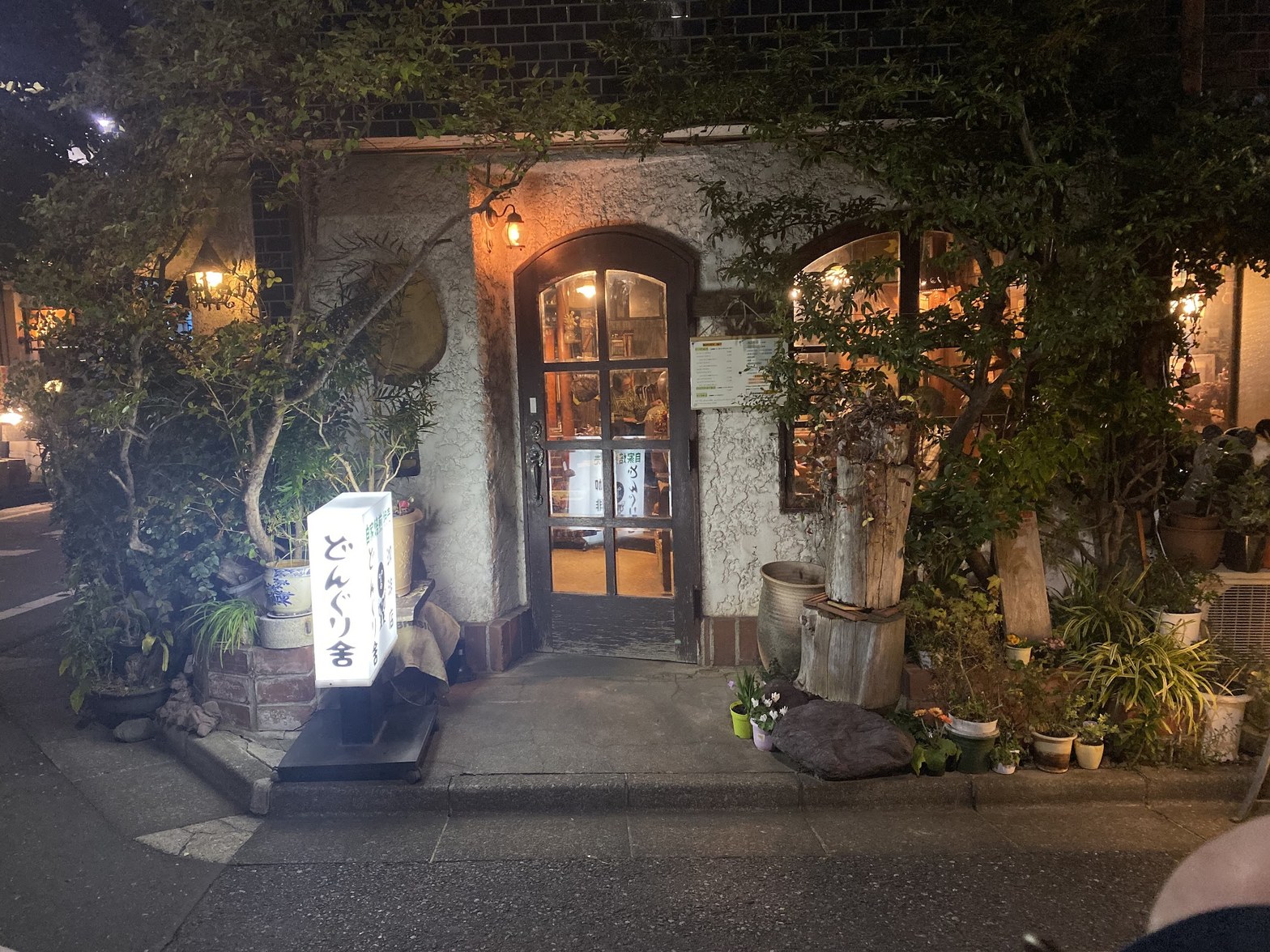 夜の外観。アーチ型ドアと植栽に囲まれた白漆喰の建物、手前に光る「どんぐり舎」の立て看板